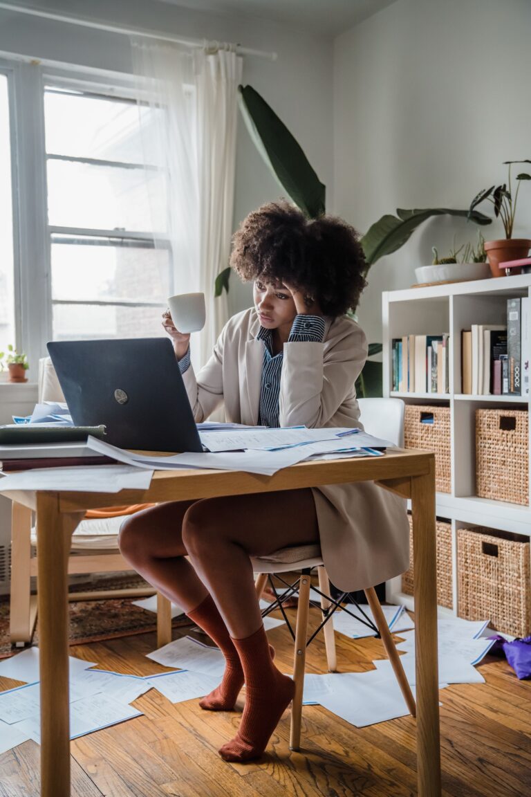 Tired woman holding a mug while looking at a laptop and paperwork at home